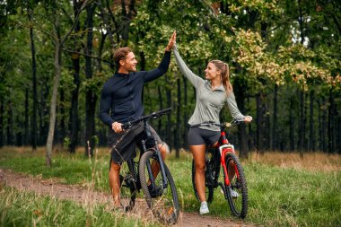 A joyful couple is biking outdoors, celebrating their adventurous spirit with highfives amid stunning greenery, enjoying life with radiant smiles and laughter, creating unforgettable memories