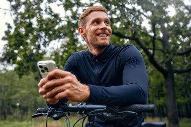 Neşeli ve hevesli bir bisikletçi açık havada keyifli bir anın tadını çıkarıyor, bisikletinin üzerinde dinlenirken mutlu bir şekilde güzel bir park manzarası seyrediyor.