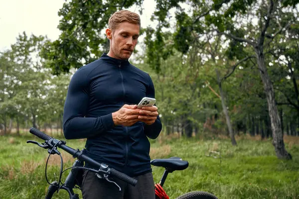 Atletizm kıyafetleri içindeki bir adam, canlı ağaçlar ve taze otlarla çevrili akıllı telefonunu dağ bisikletinin yanında kontrol ederek, keyif ve uygunluğu dengeleyen açık hava maceraları için ideal bir ortam yaratıyor.