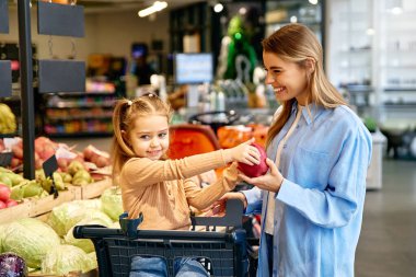 Marketin üretim bölümünde gülümseyen annesine kırmızı elma uzatan neşeli bir genç kız. Alışveriş yaparken aile içi etkileşim.