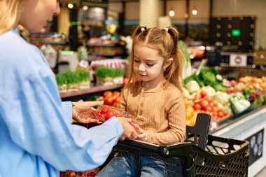 Alışveriş arabasındaki gülümseyen kız taze sebze reyonunda annesi tarafından verilen vişneli domateslere bakıyor. Tatlı günlük ebeveynlik anı.