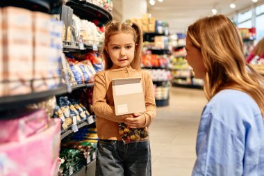 Küçük mutlu kız elinde yiyecek paketi ve süpermarket koridorunda dururken kameraya gülümsüyor. Ailemle sıcak bir alışveriş anı..