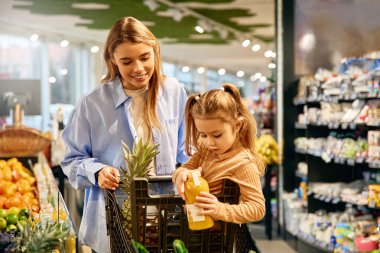 Curious young girl sitting in a shopping cart examining a juice bottle while her smiling mother stands nearby. They are grocery shopping together, creating a warm family moment.