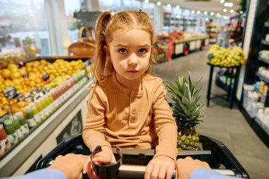 Front view of a thoughtful girl sitting in a shopping cart, holding the handlebar in a supermarket. She looks directly at the camera with a serious face.