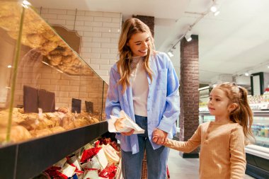 Smiling mom and little girl holding hands and standing by the bakery counter at a grocery store. The woman holding fresh bread in paper. Concept of daily shopping, family love and connection.