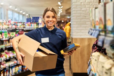 Markette çalışırken elinde karton kutu ve yiyecek olan denizci üniformalı bir kadın. Parlak, düzenli koridor. Perakende iş, lojistik ve müşteri hizmetleri kavramı.