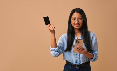 Genç bir kadın bir elinde akıllı bir telefon, diğer elinde parlak bir siyah kart tutarken güvenle gülümsüyor, yumuşak bej bir arkaplan üzerine kurulmuş, minimalist, çarpıcı bir portre yaratıyor.