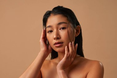 Uzun saçlı bir kadın bronz bir duvarın önünde duruyor. O, elleri yüzünde kameraya bakıyor.