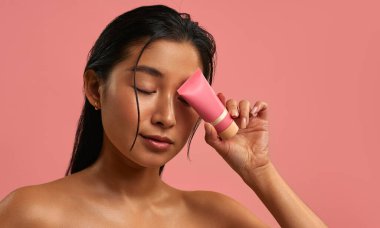 Pürüzsüz, parlak ciltli genç Asyalı kadın pembe bir kozmetik tüpü kapalı gözünün yanında tutuyor. Pastel pembe arka planda cilt bakımı. Doğal güzellik, sağlık ve yüz bakımı konsepti..
