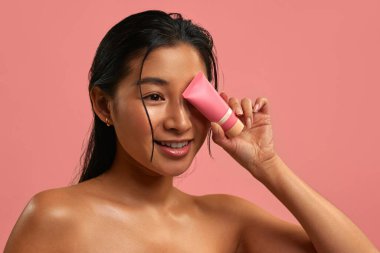 Pürüzsüz parlak ciltli genç Asyalı kadın gözünün yanında pembe bir kozmetik tüp tutuyor. Pastel pembe arka planda cilt bakımı. Doğal güzellik, sağlık ve yüz bakımı konsepti..