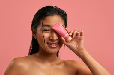 Pürüzsüz parlak ciltli genç Asyalı kadın gözünün yanında pembe bir kozmetik tüp tutuyor. Pastel pembe arka planda cilt bakımı. Doğal güzellik, sağlık ve yüz bakımı konsepti..