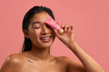 Pürüzsüz parlak ciltli genç Asyalı kadın gözünün yanında pembe bir kozmetik tüp tutuyor. Pastel pembe arka planda cilt bakımı. Doğal güzellik, sağlık ve yüz bakımı konsepti..