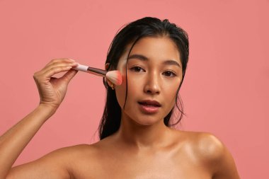 Yanağında makyaj fırçasıyla allık süren genç bir Asyalı kadın. Cilt bakımı ve pastel pembe arka planda güzellik kavramı. Doğal görünüş, parlayan ten ve sağlıklı ışık için günlük makyaj..