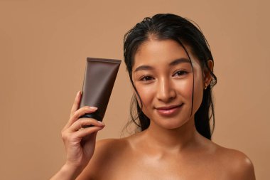 Elinde yüz kremi tutan Asyalı kadın özellikle cildinizin doğal ışıltısını arttırmak için formüle edilmiş. Bej arka planda gençleştirici izole edilmiş..