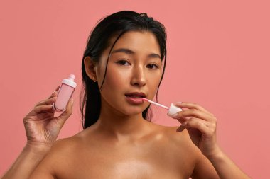 Asyalı kadın aplikatörle pembe dudak parlatıcısı sürüyor. Makyaj malzemesi pastel pembe arka planda tutuluyor. Doğal pırıl pırıl görünüş, pürüzsüz parlak cilt ve dudaklar ve yüz bakımı için güzellik rutini..