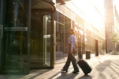 İş kıyafetli şık bir kadın bavulu çeker, güneş doğarken camdan ofis binasına girer. İş seyahati, kariyer, başarı ve şehir yaşam tarzı kavramı.