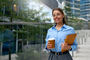 Mavi gömlekli ve gözlüklü, gülümseyen, elinde tablet ve kahve fincanı tutan genç profesyonel bir kadın modern şehir ortamında. Güven, başarı ve iş hayatı dengesi.
