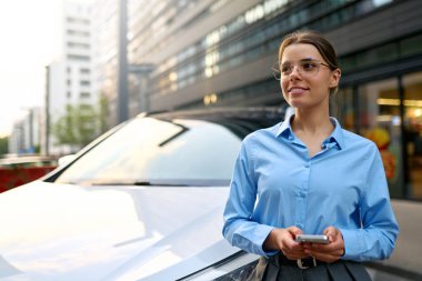 Şehirde beyaz bir arabanın yanında akıllı telefon kullanırken gülümseyen mavi gömlekli genç bir iş kadını. Modern yaşam tarzı, şehir manzarası, iş gezisi konsepti..