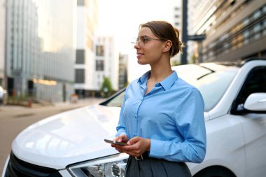 Şehirde beyaz bir arabanın yanında akıllı telefon kullanırken gülümseyen mavi gömlekli genç bir iş kadını. Modern yaşam tarzı, şehir manzarası, iş gezisi konsepti..
