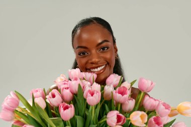 Elinde pembe lale buketi tutan Afro-Amerikalı kadın Anneler Günü 'nü ya da Kadınlar Günü' nü neşe ve sevgiyle kutluyor. Tarafsız bir arkaplanda gülümsüyor. Bahar tatili ve kadınsı güzellik kavramı.