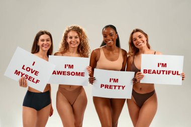 Farklı etnik kökenlere sahip dört gülümseyen kadın. Onaylı pankartlar taşımaktan gurur duyuyorlar. Kendini sevme, güçlendirme ve beden kabulü kavramı.
