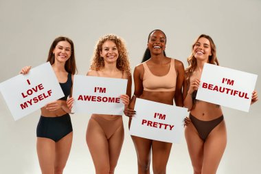 Farklı etnik kökenlere sahip dört gülümseyen kadın. Onaylı pankartlar taşımaktan gurur duyuyorlar. Kendini sevme, güçlendirme ve beden kabulü kavramı.
