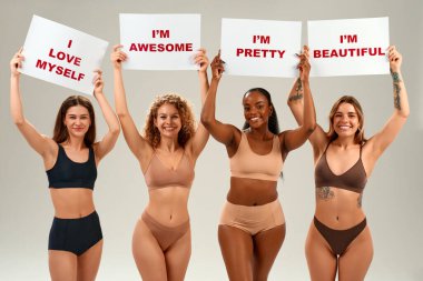 Farklı etnik kökenlere sahip dört gülümseyen kadın. Onaylı pankartlar taşımaktan gurur duyuyorlar. Kendini sevme, güçlendirme ve beden kabulü kavramı.