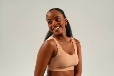 Çıplak spor bluzuyla genç Afrikalı bir kadın, kameraya parlak ve kendinden emin bir şekilde gülümsüyor, cilt bakımını, güzelliği, kozmetolojiyi, sağlık konseptini temsil ediyor..