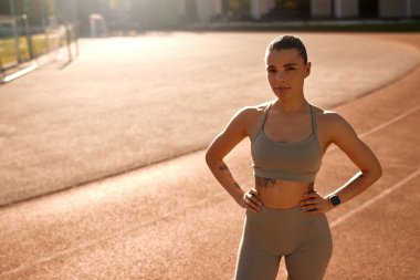 Güçlü atletik kadın, spor kıyafetleriyle stadyum pistinde duruyor, elleri kalçasında, kendinden emin görünüyor. Konsept: Kadın gücü, spor, sağlıklı yaşam tarzı, kendine güven, sabah sporu.