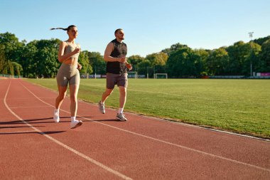Genç ve zinde bir çift sabahleyin stadyum pistinde birlikte koşuyor. Sağlıklı yaşam tarzı, motivasyon ve güneş ışığı rutin konsepti.