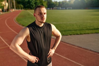 Spor yaptıktan sonra elleri kalçasında koşu pistinde duran kaslı genç adamın portresi. Disiplin, güç ve atletik yaşam tarzı kavramı.