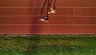 Atletik erkek ve kadının çimlerin yakınındaki kırmızı bir pistte yan yana koştuğunu gösteren drone üst görüntüsü. Uzun gölgeler, akşam güneşi. Spor anlayışı, dayanıklılık ve sağlıklı yaşam tarzı.