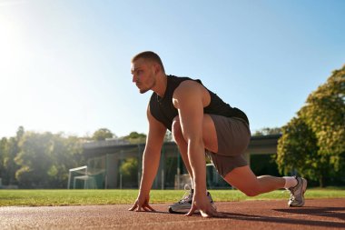 Formda genç bir adam gün doğumunda açık hava pistinde koşmaya hazırlanıyor. Odaklanmış ve kararlı ifade. Disiplin, hız ve sağlıklı yaşam tarzı kavramı.