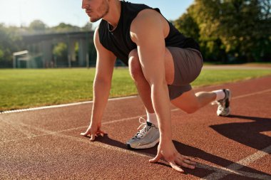 Formda genç bir adam gün doğumunda açık hava pistinde koşmaya hazırlanıyor. Odaklanmış ve kararlı ifade. Disiplin, hız ve sağlıklı yaşam tarzı kavramı.