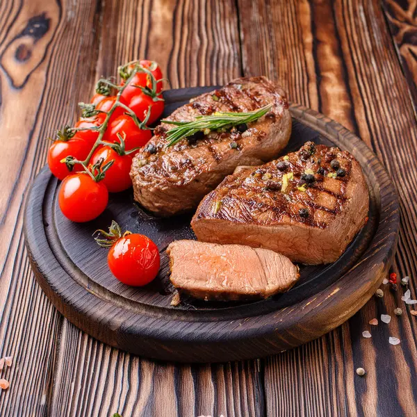 Juicy medium Beef Rib Eye steak slices in pan on wooden board with fork and knife herbs spices and salt.