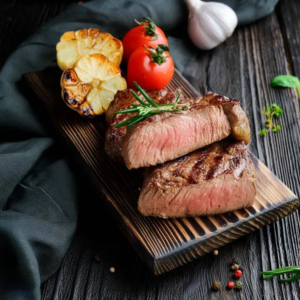 Juicy medium Beef Rib Eye steak slices in pan on wooden board with fork and knife herbs spices and salt.