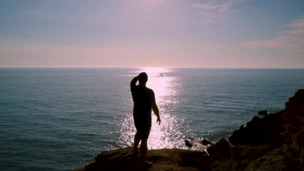 背景の海の水に太陽の反射の軌跡と崖の上の男のシルエットは 概念を作る — ストック動画
