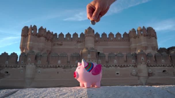 在阳光明媚的日子里 一只手把钱放进一个有着中世纪城堡的独角兽小猪银行 背景是蓝天蓝云 — 图库视频影像