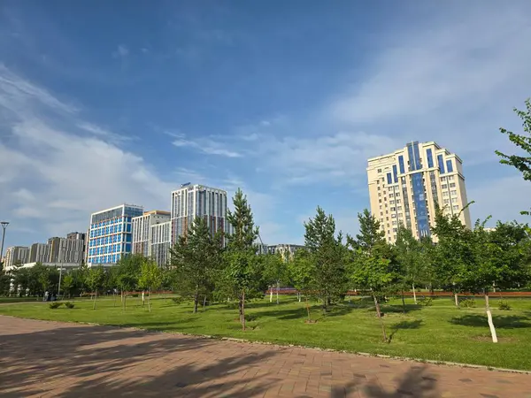 Asfaltlı bir yürüyüş yolu, yemyeşil çimenler ve modern gökyüzü altındaki yüksek binalar tarafından çerçevelenmiş genç ağaçlarla dolu manzaralı bir şehir parkı, şehir ve doğa tasarımları için ideal..