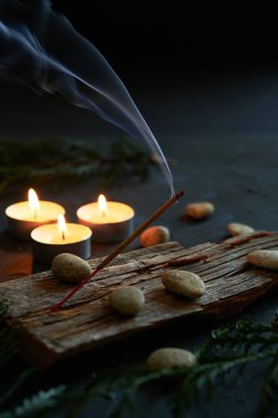 Tütsü çubuğundan çıkan yumuşak bir duman odayı tatlı ve topraksı bir kokuyla dolduruyor. Arka plandaki küçük mumlar, huzurlu ve rahatlatıcı bir ortam yaratmak için ılık ve nazik bir ışık yayıyor.