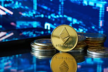 Tek bir Ethereum madeni parası, arkasında başka paralar olan bir masanın üzerinde duruyor. Arka planda mali verileri olan bulanık bir bilgisayar ekranı var.