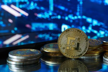 Tek bir Binance madeni parası, arkasında başka paralar olan bir masanın üzerinde duruyor. Arka planda mali verileri olan bulanık bir bilgisayar ekranı var.