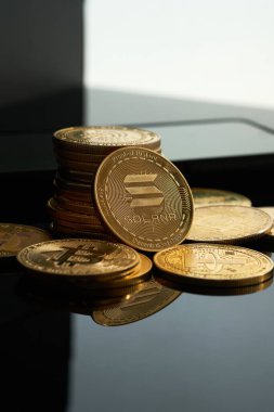 Bir yığın altın Solana sikkesi siyah bir yüzeye yayılmış ve diğer paralar etrafa saçılmıştır. Arka planda bir akıllı telefon görebilirsiniz..
