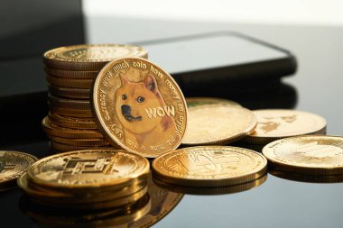 Bir yığın altın Dogecoin sikkesi siyah bir yüzeye yayılmış ve diğer madeni paralar etrafa saçılmıştır. Arka planda bir akıllı telefon görebilirsiniz..