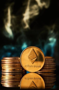 Parlak bir yüzey üzerinde belirgin bir şekilde gösterilen Ethereum sikkeleri yığını, kripto para temasını geliştiren dumanlı ve renkli bir arka plan.