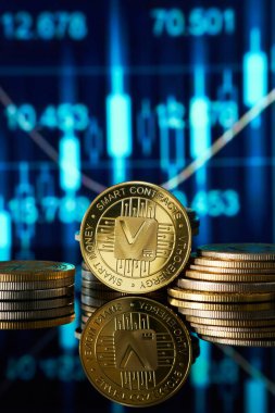 Stacks of golden Vethor cryptocurrency coins are reflected on a smooth surface, with a vibrant digital trading chart showing market trends in the background, highlighting a tech-savvy financial environment. clipart