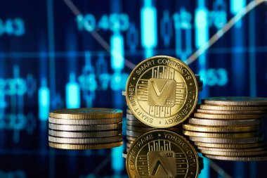 Altın Vethor kripto para yığınları pürüzsüz bir yüzeye yansıtılır, arka planda piyasa eğilimlerini gösteren canlı dijital ticaret grafiği teknolojiden anlayan finansal ortamı vurgular..