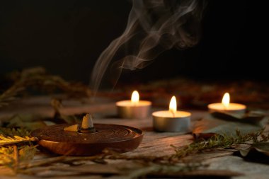 Tütsü dumanının yumuşak kıvrımları, titreyen mum alevleriyle birlikte ahşap bir tutacaktan yükselir. Yapraklar düzenlemeyi sarar, sakinleştirici titreşimleri arttırır.