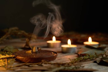 Tütsü dumanının yumuşak kıvrımları, titreyen mum alevleriyle birlikte ahşap bir tutacaktan yükselir. Yapraklar düzenlemeyi sarar, sakinleştirici titreşimleri arttırır.