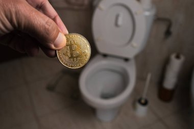 Bir el, tuvaletin üstünde parlak bir Bitcoin para sunar. Banyo kiremitleri, tuvalet kağıtları ve tuvalet malzemeleri kripto para birimiyle komik bir kontrast oluşturuyor..